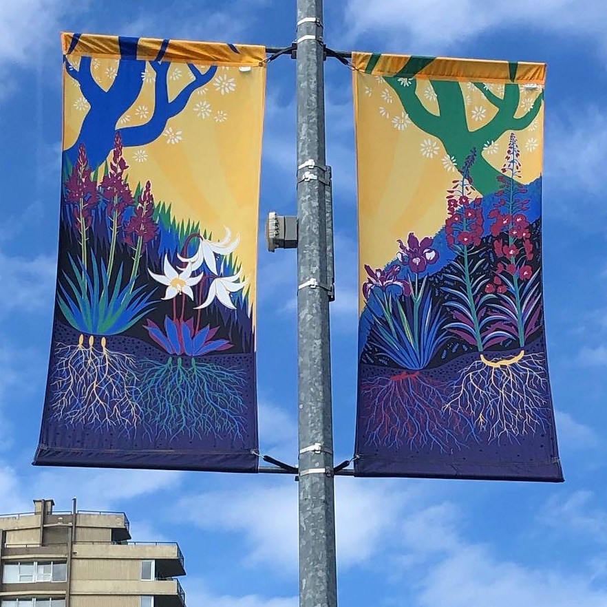 street banner hanging from lamp post, in two panels against a blue and clouded sky. Background colour is yellow, with a colourful scene of four flowers rooted into the ground, against grass and berry bushes, with tree branches hanging above.