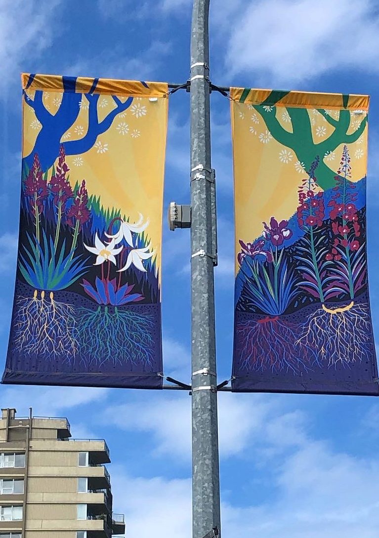 street banner hanging from lamp post, in two panels against a blue and clouded sky. Background colour is yellow, with a colourful scene of four flowers rooted into the ground, against grass and berry bushes, with tree branches hanging above.
